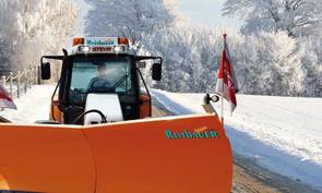 Auf dem Bild ist ein Traktor mit Schneepflug in winterlicher Landschaft zu sehen.